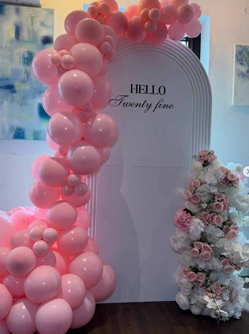 Pink balloon garland around a white arch board reading Hello Twenty Fine with floral column