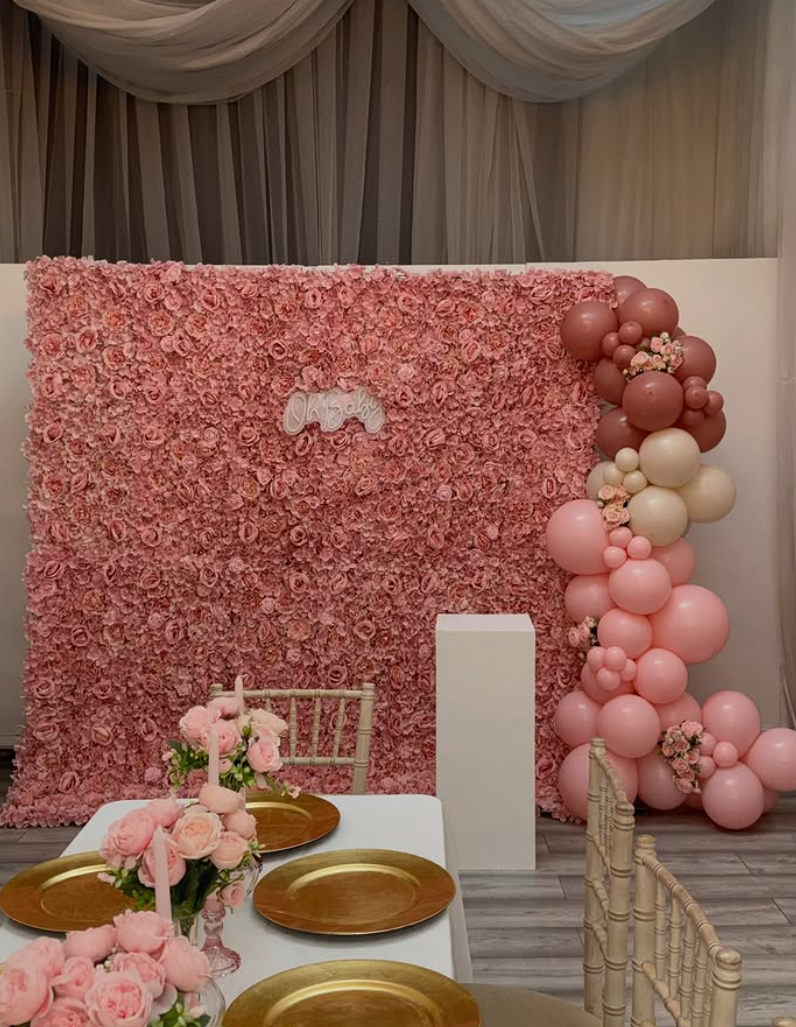 Pink flower wall with an Oh Baby neon sign and a pink balloon garland beside a styled table